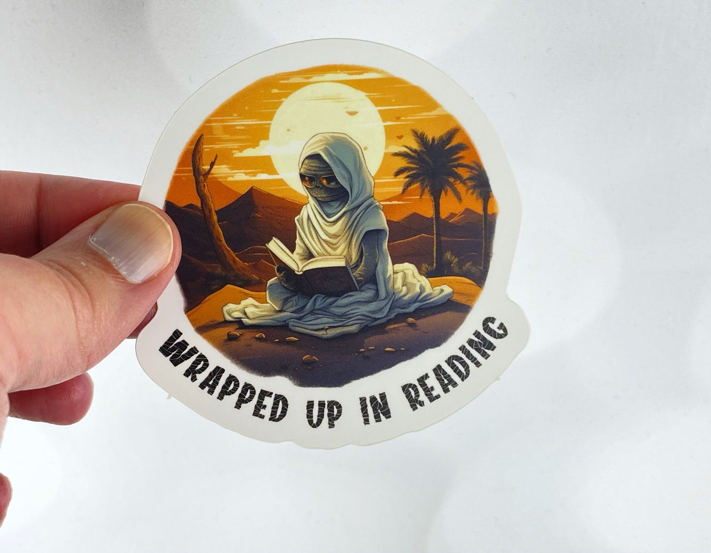 A hand holds a sticker illustration of a hooded desert figure reading a book under a moon with palm trees in the background, reading "Wrapped up in reading," against a white background.