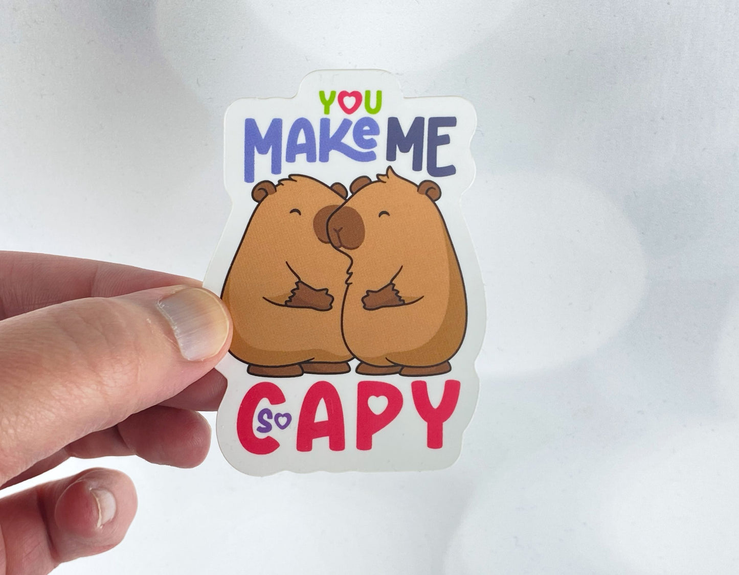 Die-cut vinyl sticker held between fingers, showing two capybaras nuzzling each other, above the text "You make me so capy," on a white background