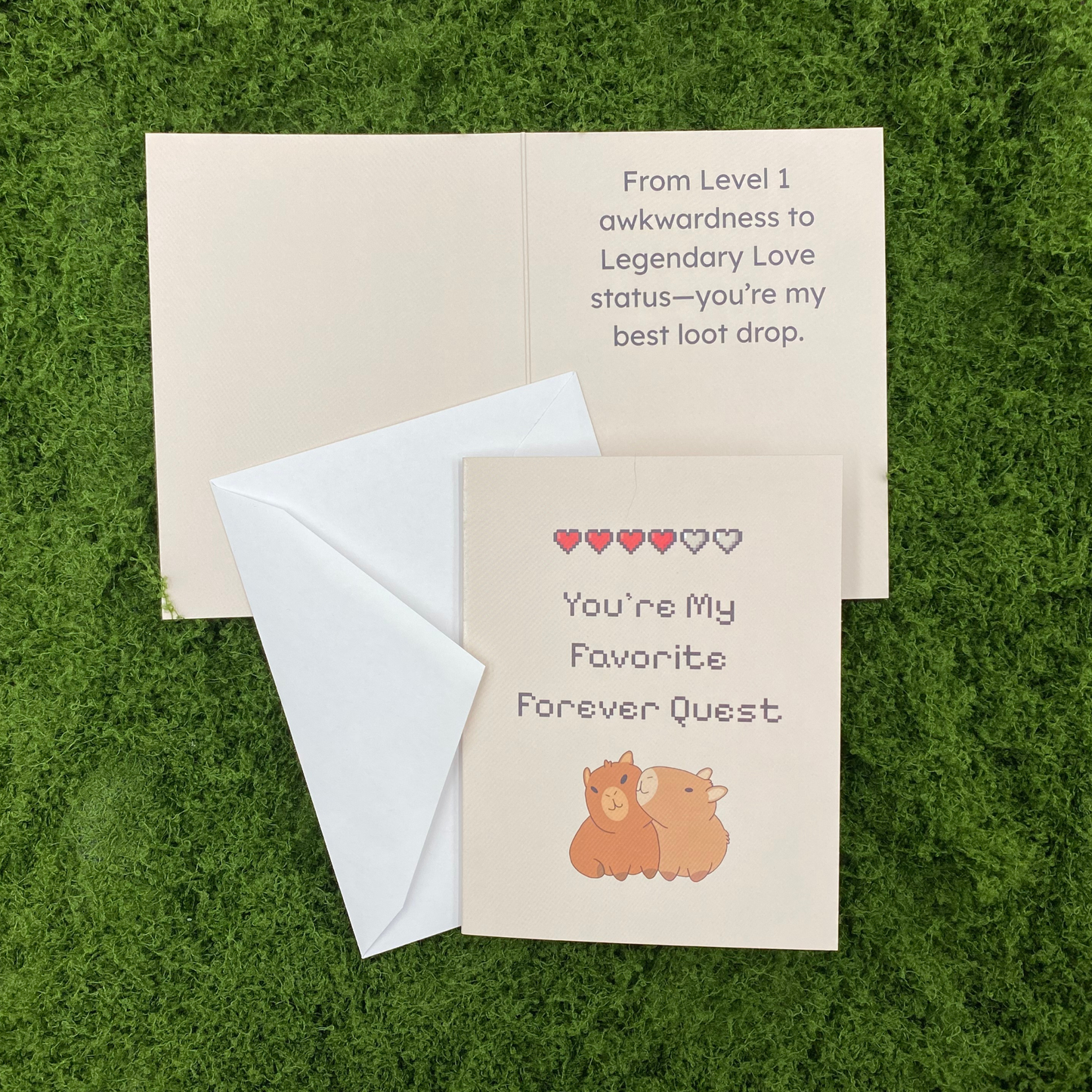 Greeting card lying open on artificial grass, showing the front with a pixel-art heart meter and "You're My Favorite Forever Quest" above an illustration of two cuddling capybaras, next to the inside message "From Level 1 awkwardness to Legendary Love status—you're my best loot drop," with the envelope tucked beneath the front cover.