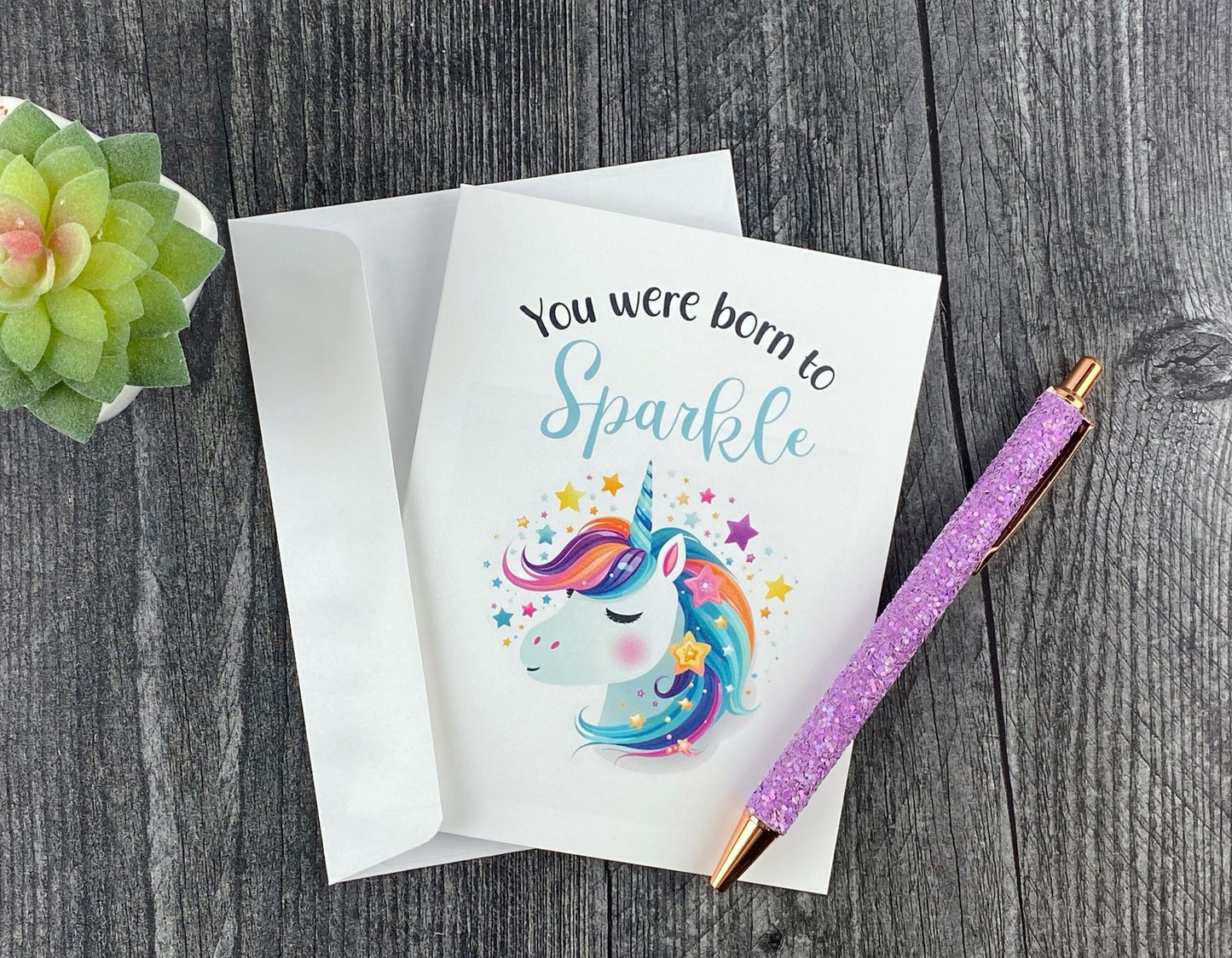 Greeting card and white envelope, featuring an illustrated rainbow-maned unicorn surrounded by stars, reading "You were born to sparkle," resting on a grey wood surface next to a purple glitter pen and small succulent.