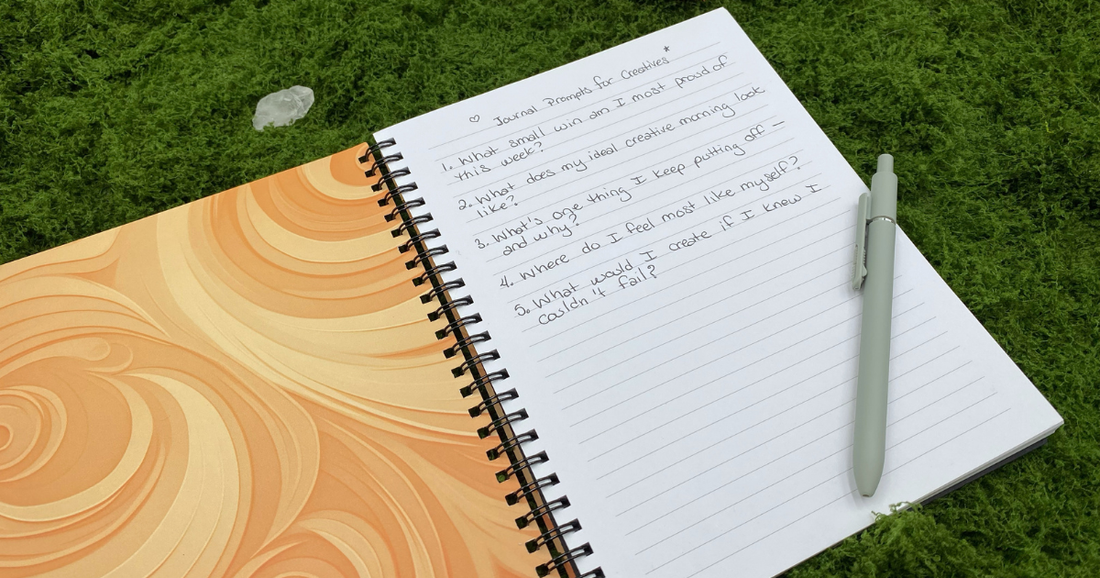 Wire-o notebook open to a handwritten journaling prompts page on a green moss mat, with the orange swirl cover visible on the left, a light green pen resting on the right page, and a clear quartz crystal in the upper left corner