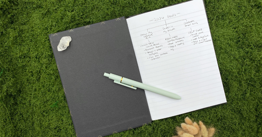 Hardcover journal open to a handwritten 2026 goals page on a green moss mat, styled with a clear quartz crystal, light green pen, and dried bunny tail grass bouquet