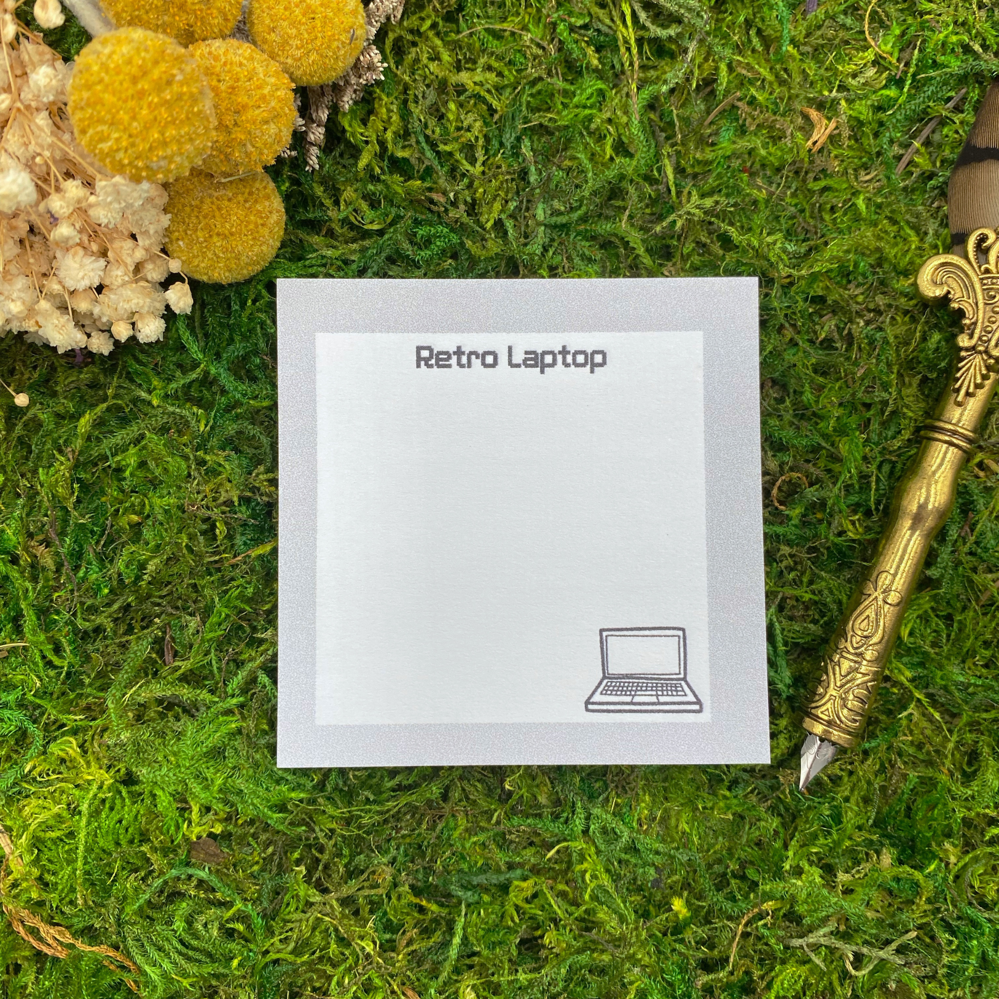 Square sticky note pad with a silver border, a pixel-font "Retro Laptop" heading, and a small hand-drawn laptop illustration, resting on moss next to dried yellow and white flowers and a gold quill pen.