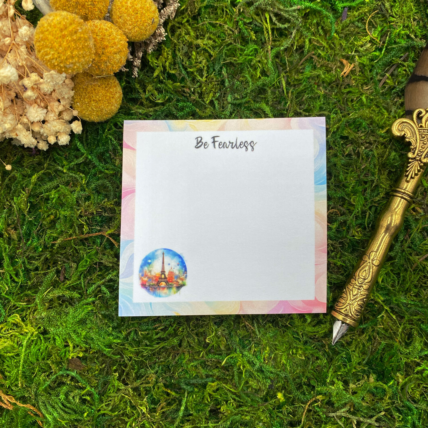 3x3 inch sticky note pad with "Be Fearless" text and watercolor Eiffel Tower illustration on pastel pink, blue, and gold border, displayed on green moss with yellow dried flowers and vintage gold fountain pen