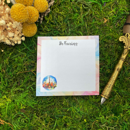 3x3 inch sticky note pad with "Be Fearless" text and watercolor Eiffel Tower illustration on pastel pink, blue, and gold border, displayed on green moss with yellow dried flowers and vintage gold fountain pen