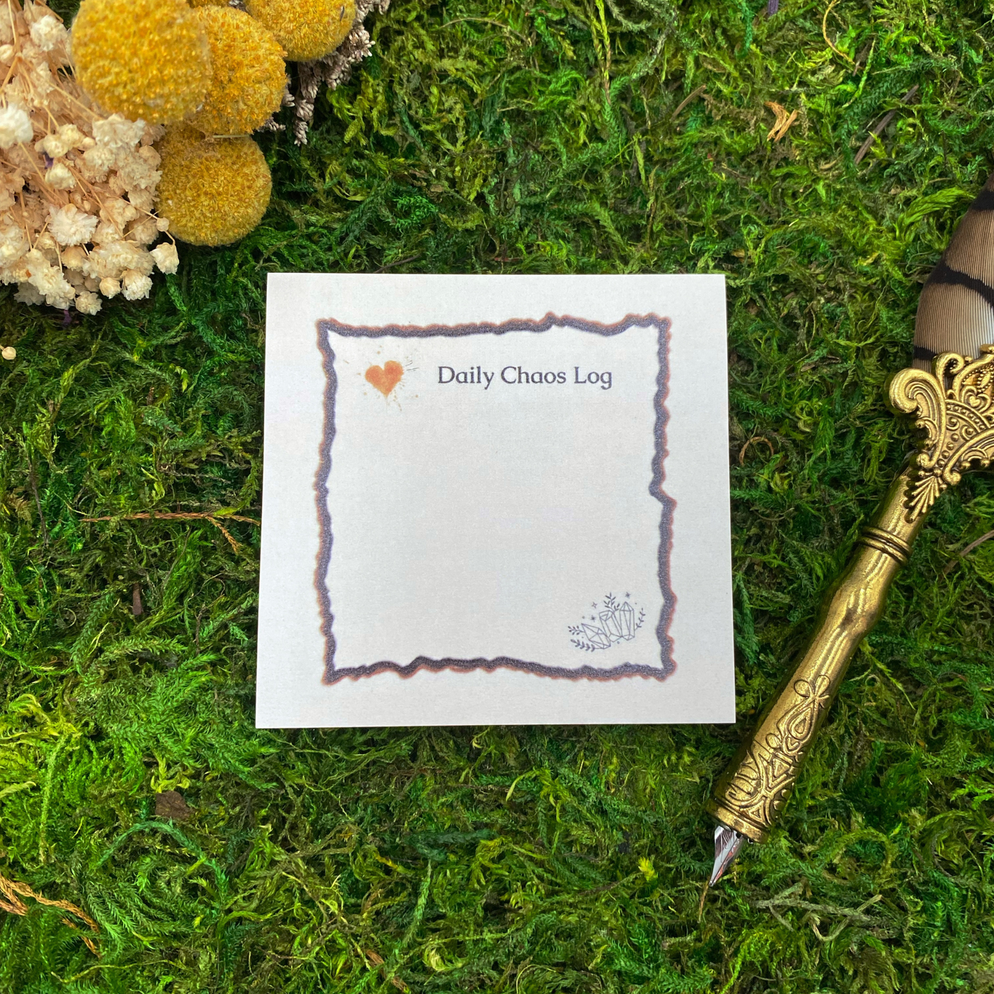 3x3 inch sticky note pad reading "Daily Chaos Log" with coffee stain heart, crystal illustrations, and hand-drawn border, displayed on green moss with yellow dried flowers and vintage gold quill pen