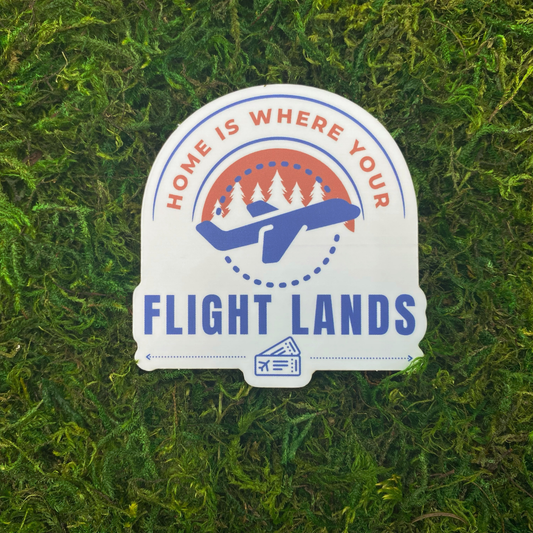 3 inch die-cut vinyl sticker with vintage airplane badge design reading "Home Is Where Your Flight Lands" featuring plane, pine trees, and boarding pass in coral and blue colors, displayed on green moss background