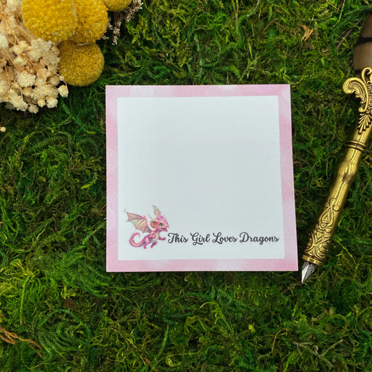 3x3 pink dragon sticky note pad reading “This Girl Loves Dragons” with a cute baby dragon illustration, styled on green moss with decorative pen.