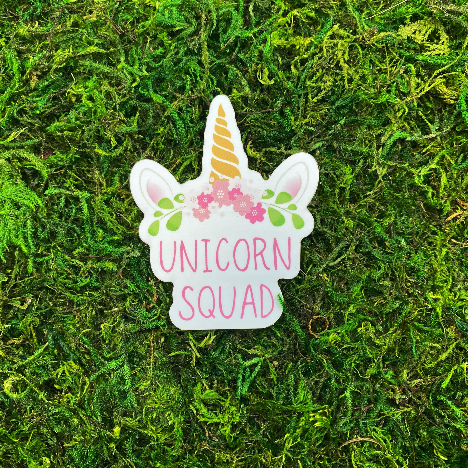 Die-cut vinyl sticker featuring a white unicorn face with a gold swirled horn, pink floral crown, and the words "Unicorn Squad" in pink lettering, photographed on a bed of green moss.