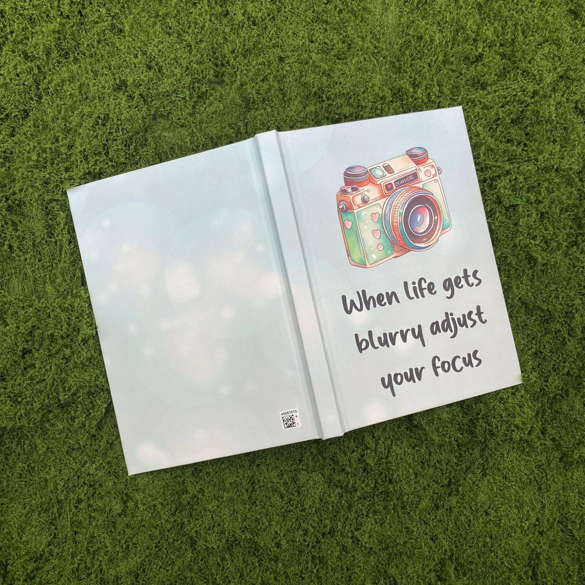 Open light blue hardcover journal showing the plain back cover and the watercolor camera design with the focus quote on the front cover.