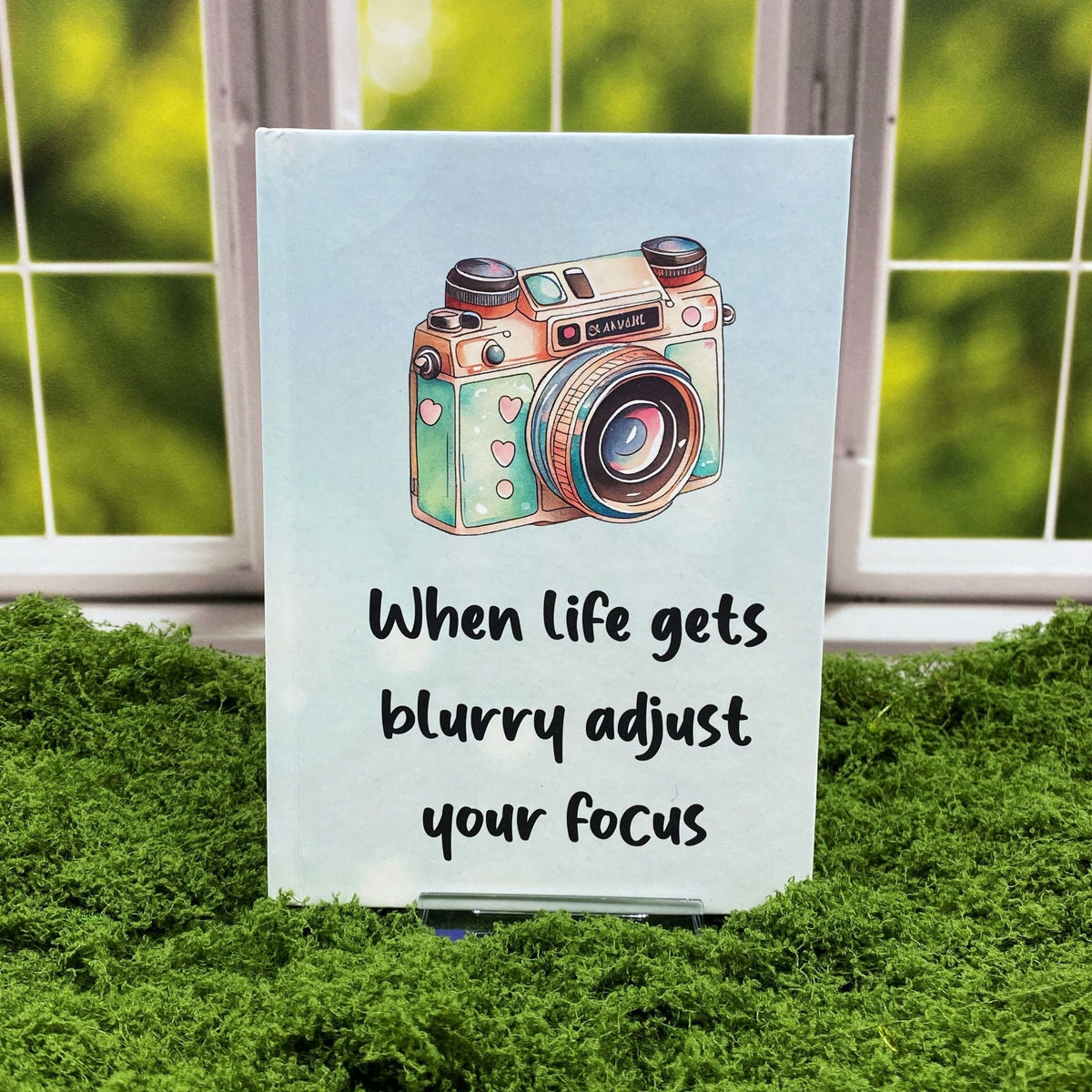 Light blue hardcover journal with a watercolor camera illustration, reading 'When life gets blurry, adjust your focus,' displayed on a moss mat with a window backdrop