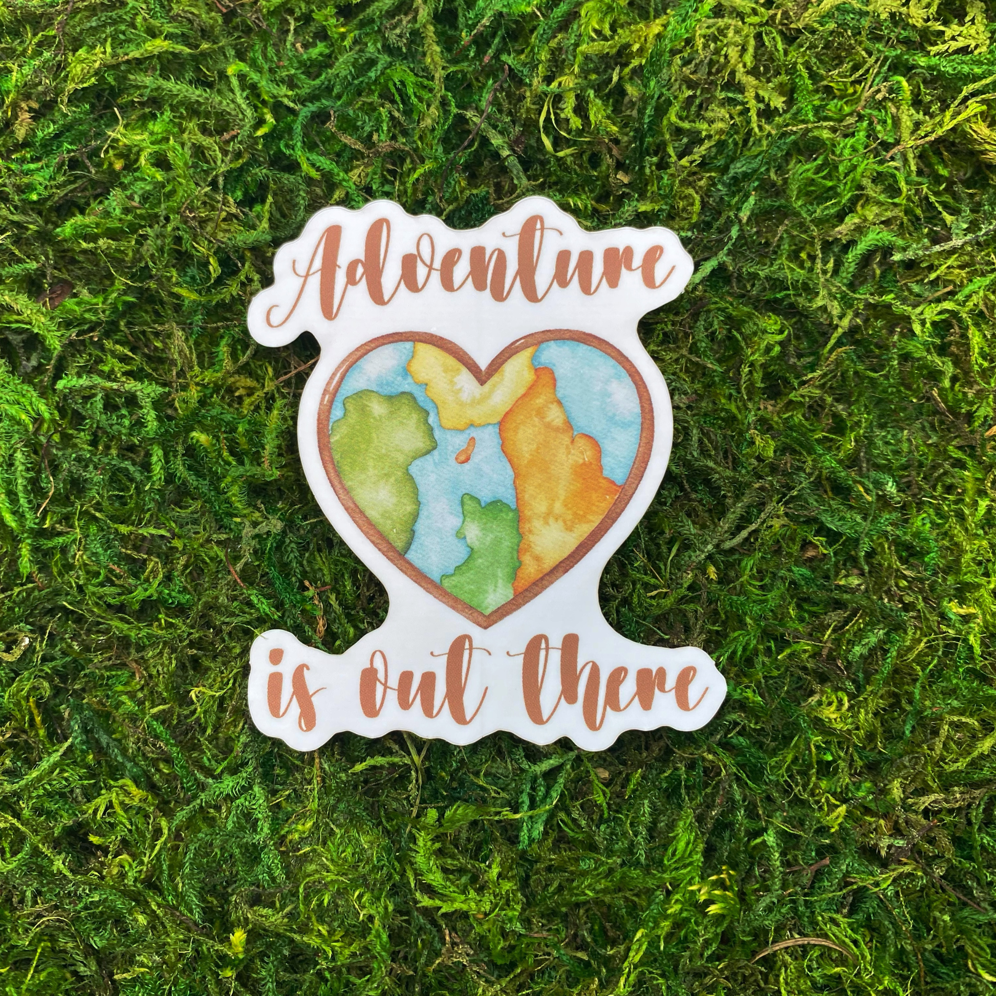 A sticker illustration of a watercolor world map shaped into a heart, reading "Adventure is out there," resting on artificial moss.
