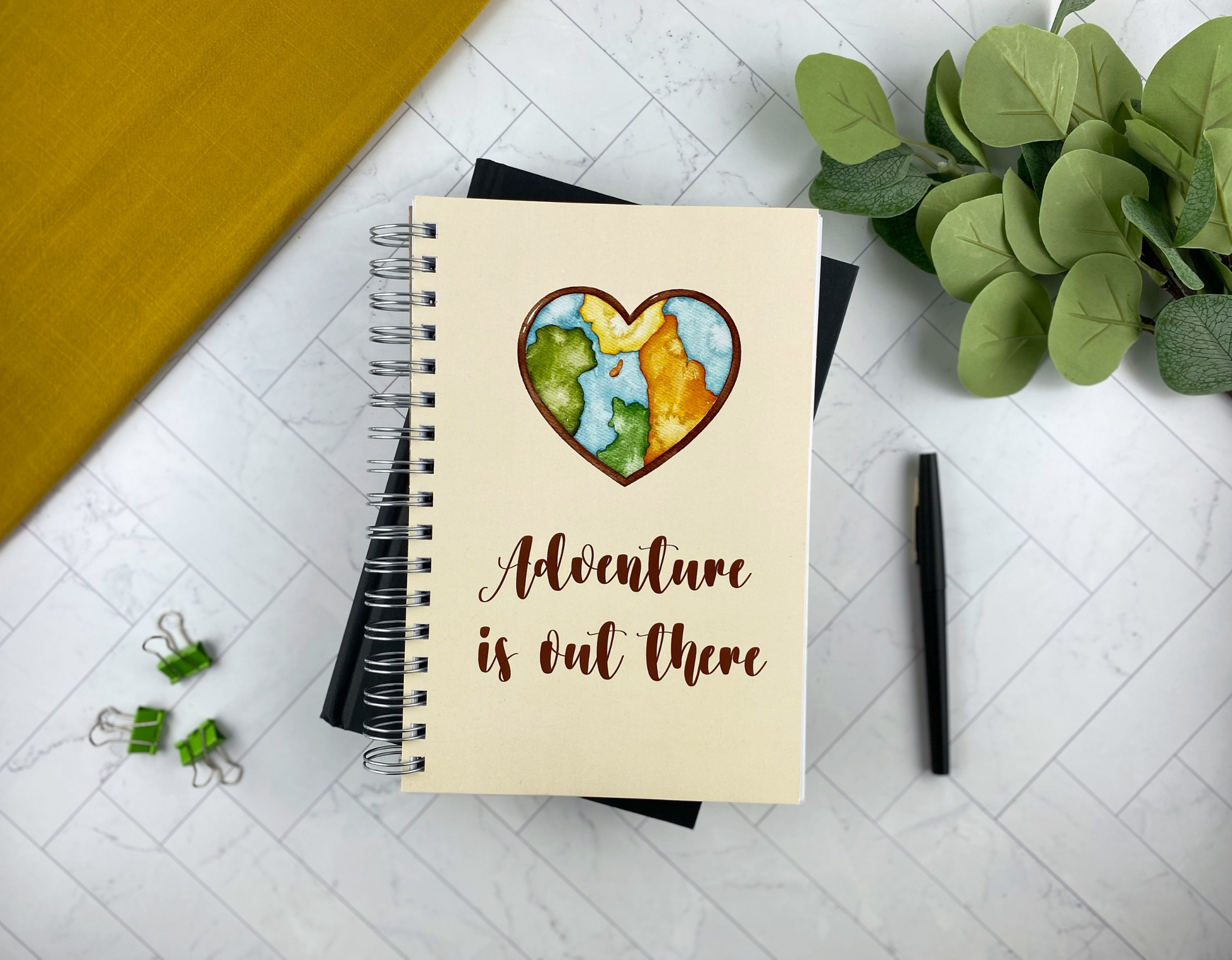 Spiral notebook cover in cream with a watercolor heart-shaped illustration of the world map and the words "Adventure is out there," resting on a black notebook with green binder clips, a black pen, and eucalyptus greenery nearby.