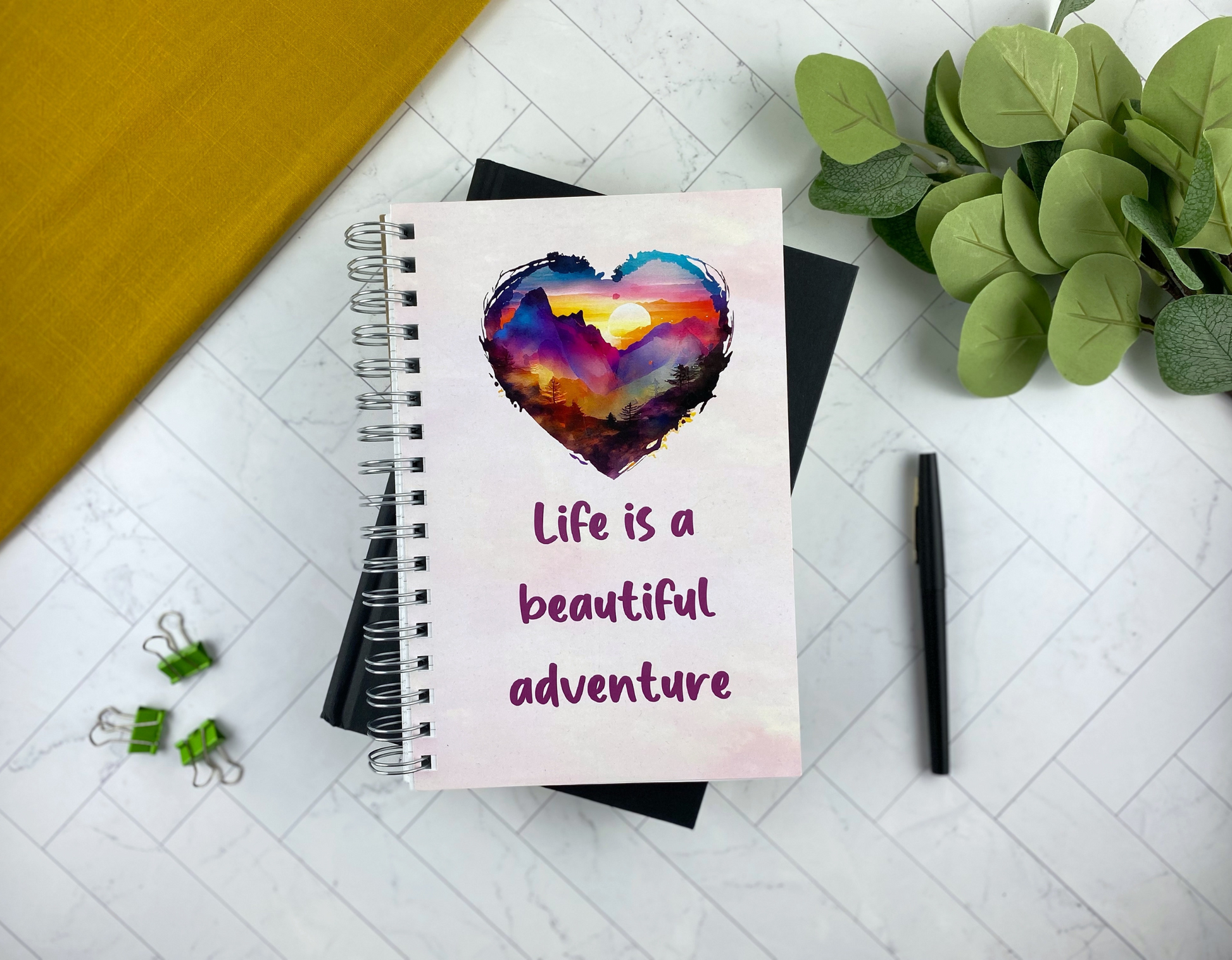 Spiral notebook cover with a heart-shaped watercolor sunset over mountains and the words "Life is a beautiful adventure," resting on a black notebook with green binder clips, a black pen, and eucalyptus greenery nearby.
