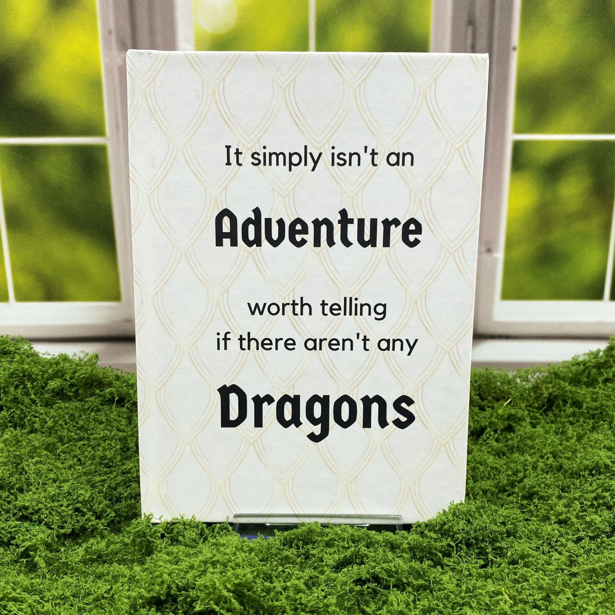 Cream hardcover journal with a dragon-scale pattern reading 'It simply isn't an adventure worth telling if there aren't any dragons,' displayed on a moss mat with a window backdrop.