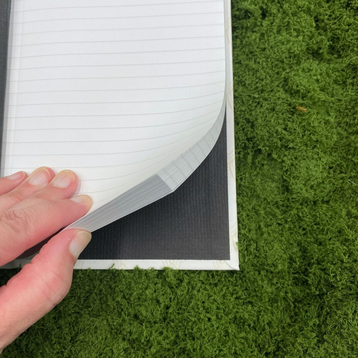 Close-up of a hand fanning through the lined pages of the journal, showing the black cardboard interior of the hardcover binding