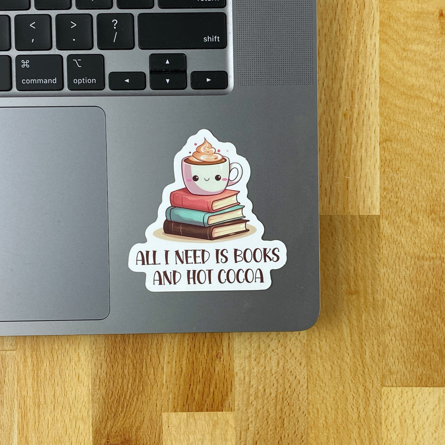 A sticker illustration of a smiling cocoa mug topped with whipped cream on a stack of three books, reading "All I need is books and hot cocoa," applied to the corner of a laptop's trackpad and keyboard area, resting on a wood desk.