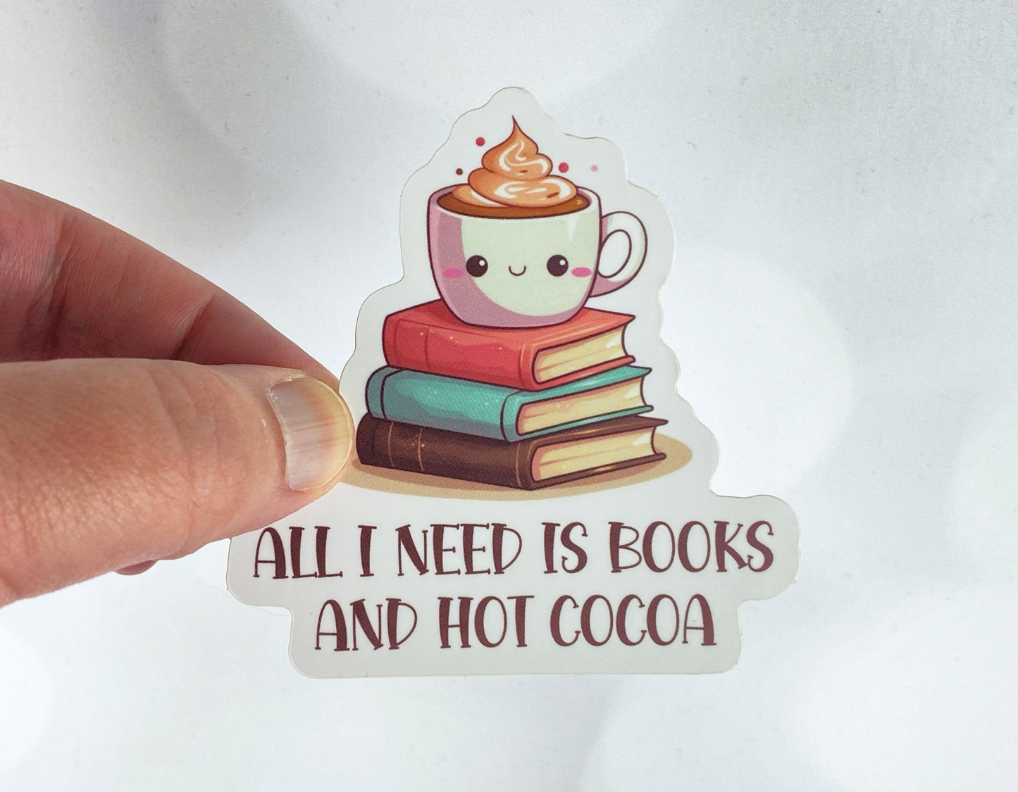 A hand holds a sticker illustration of a smiling cocoa mug topped with whipped cream, sitting on a stack of three books, reading "All I need is books and hot cocoa," against a white background.