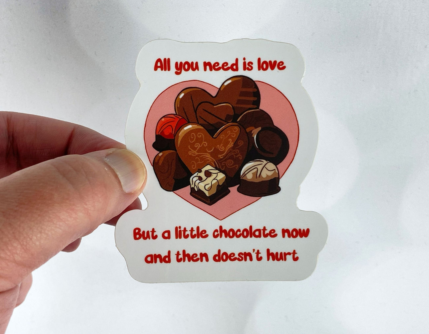 A hand holding a die-cut sticker featuring assorted chocolates in a pink heart reading all you need is love but a little chocolate now and then doesn't hurt, against a white background