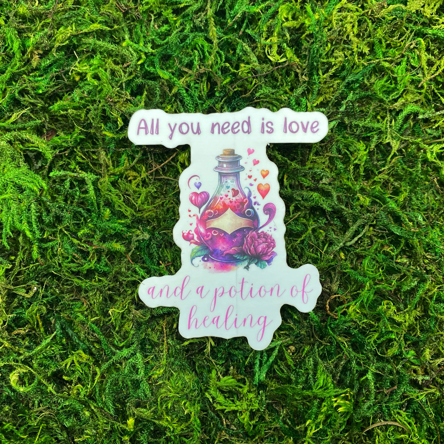 A sticker with a watercolor illustration of a potion bottle surrounded by hearts and a pink rose, reading "All you need is love and a potion of healing," resting on artificial moss.