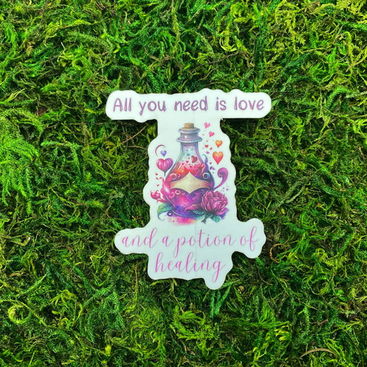 A sticker with a watercolor illustration of a potion bottle surrounded by hearts and a pink rose, reading "All you need is love and a potion of healing," resting on artificial moss.