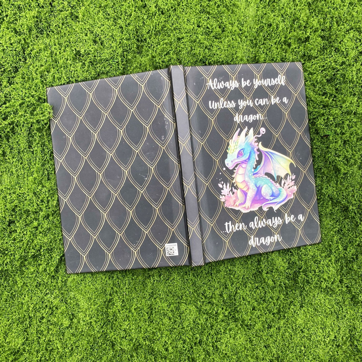 Open black hardcover journal showing the plain gold dragon-scale pattern on the back cover and the pastel baby dragon design on the front cover.