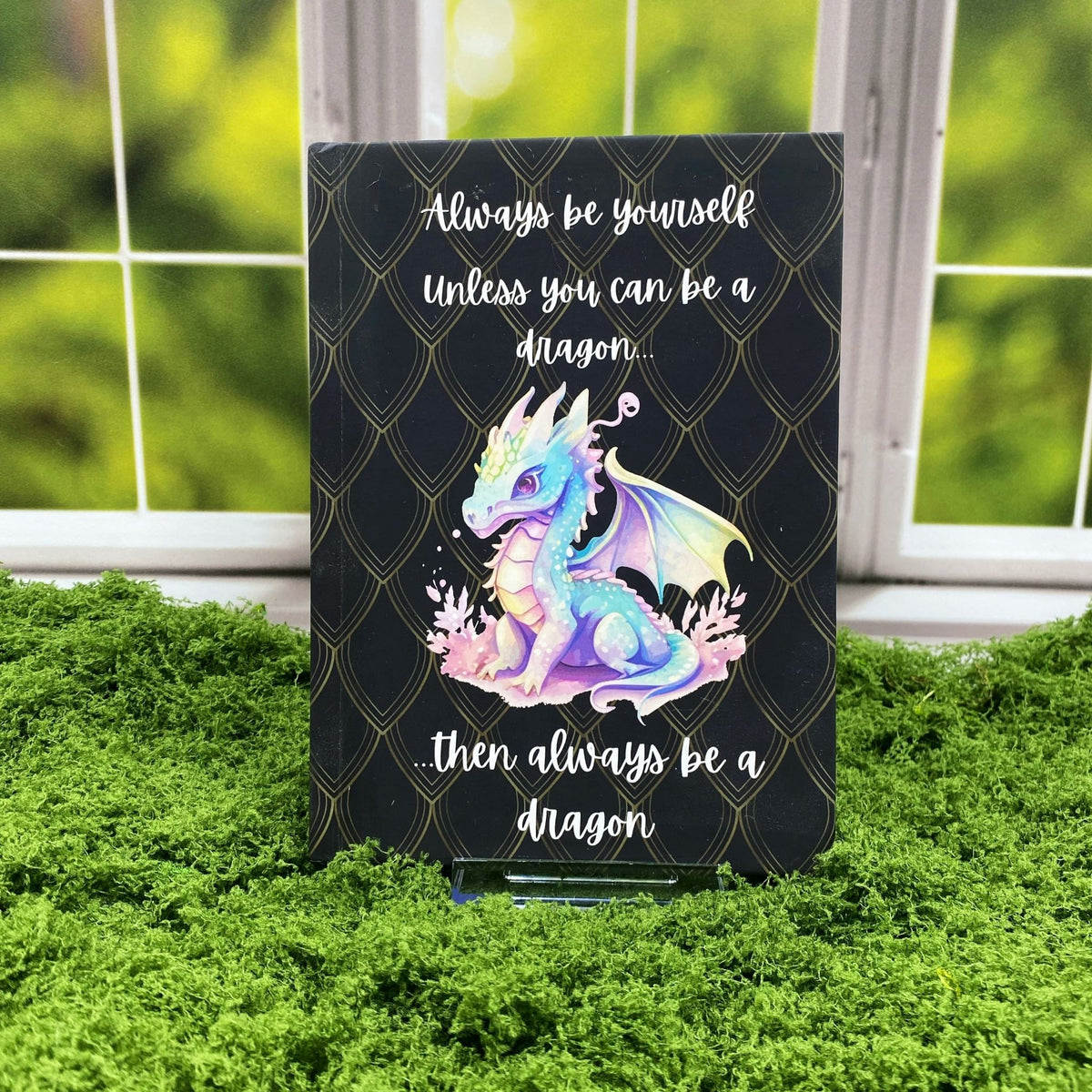 Black hardcover journal with a gold dragon-scale pattern and a pastel baby dragon illustration, reading 'Always be yourself. Unless you can be a dragon... then always be a dragon,' displayed on a moss mat with a window backdrop.