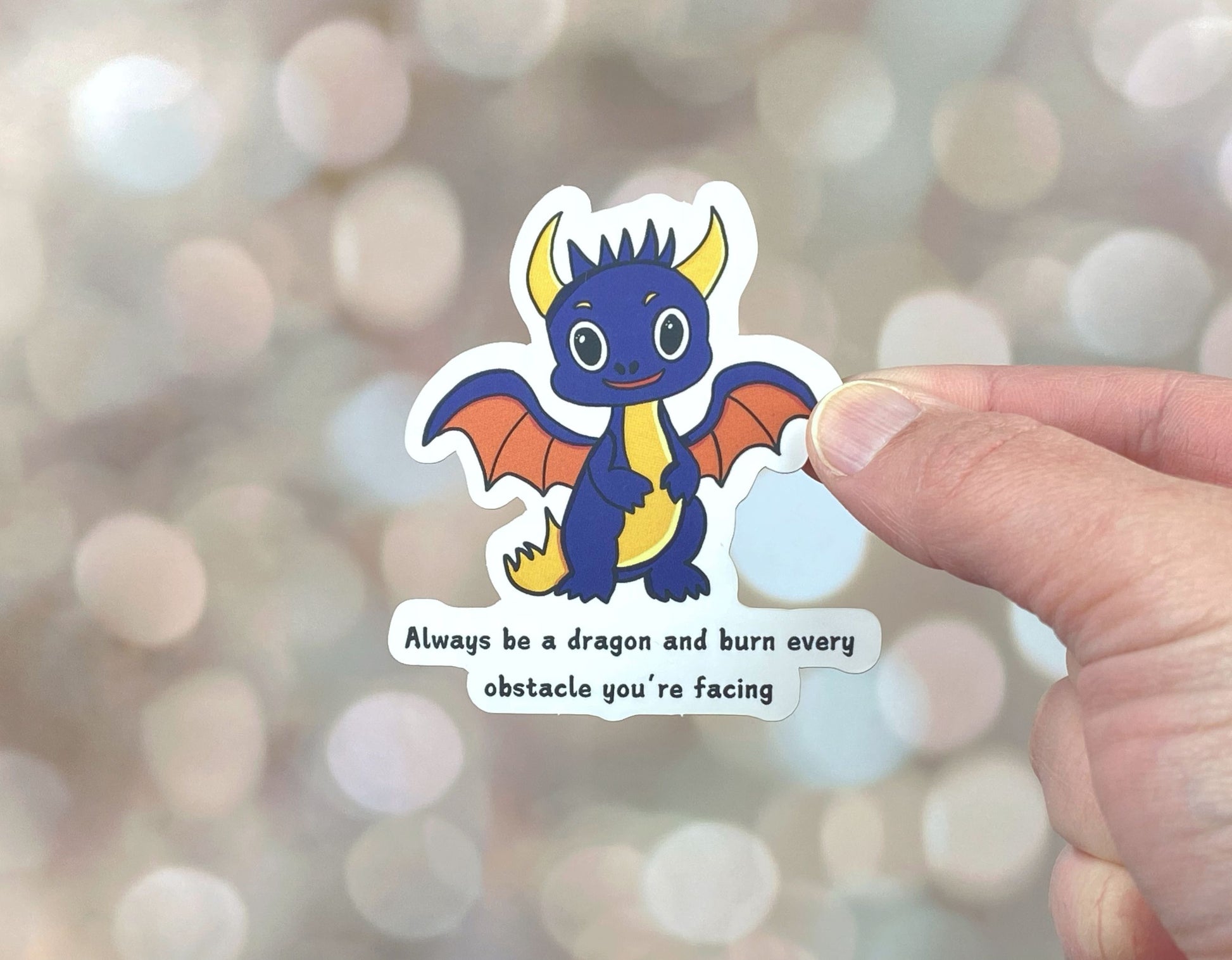 Die-cut vinyl sticker showing a dark grey baby dragon with orange wings, with the text "Always be a dragon and burn every obstacle you're facing," on a golden bokeh background