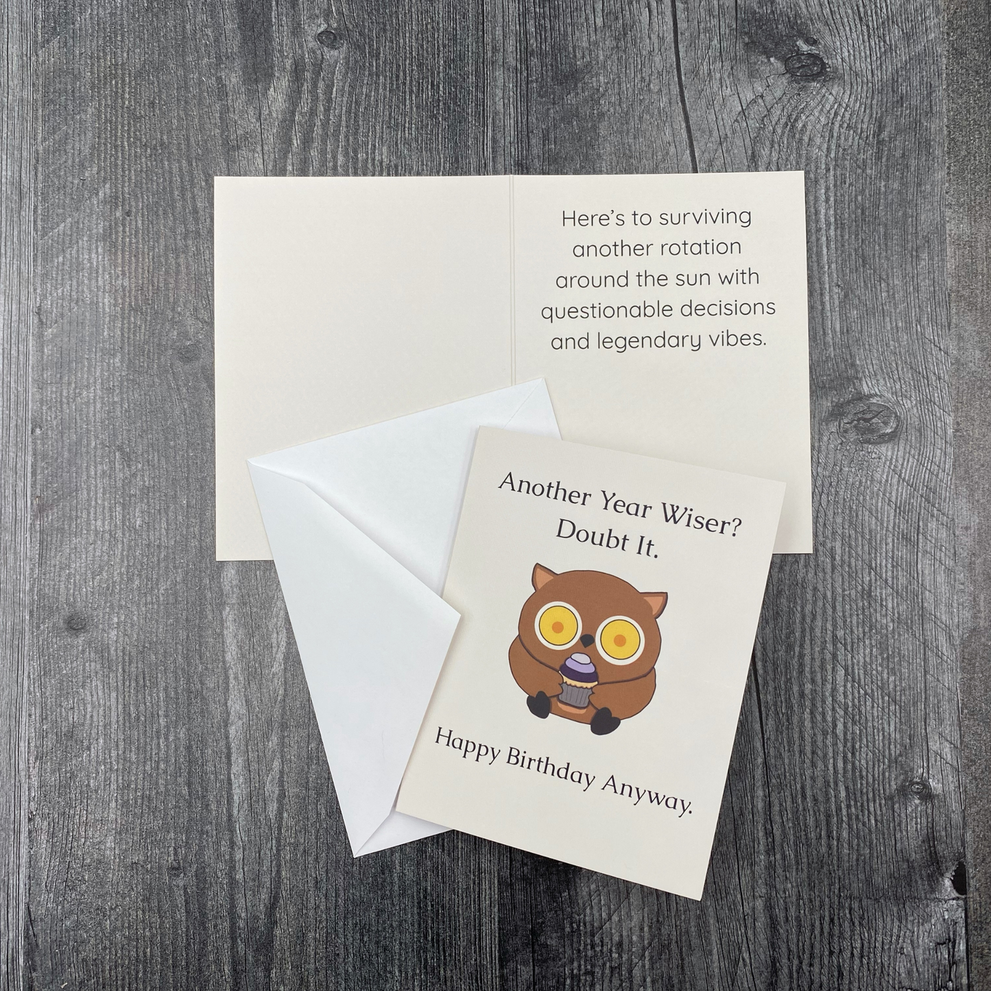 Greeting card lying open on a grey wood surface, showing the front with "Another Year Wiser? Doubt It." and an illustration of a wide-eyed owl holding a cupcake above "Happy Birthday Anyway," next to the inside message "Here's to surviving another rotation around the sun with questionable decisions and legendary vibes," with the envelope tucked beneath the front cover.