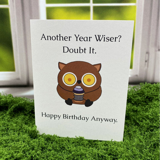 "Another Year Wiser? Doubt It. Happy Birthday Anyway." greeting card standing upright on a moss mat in front of a window backdrop, featuring an illustrated wide-eyed owl holding a cupcake.