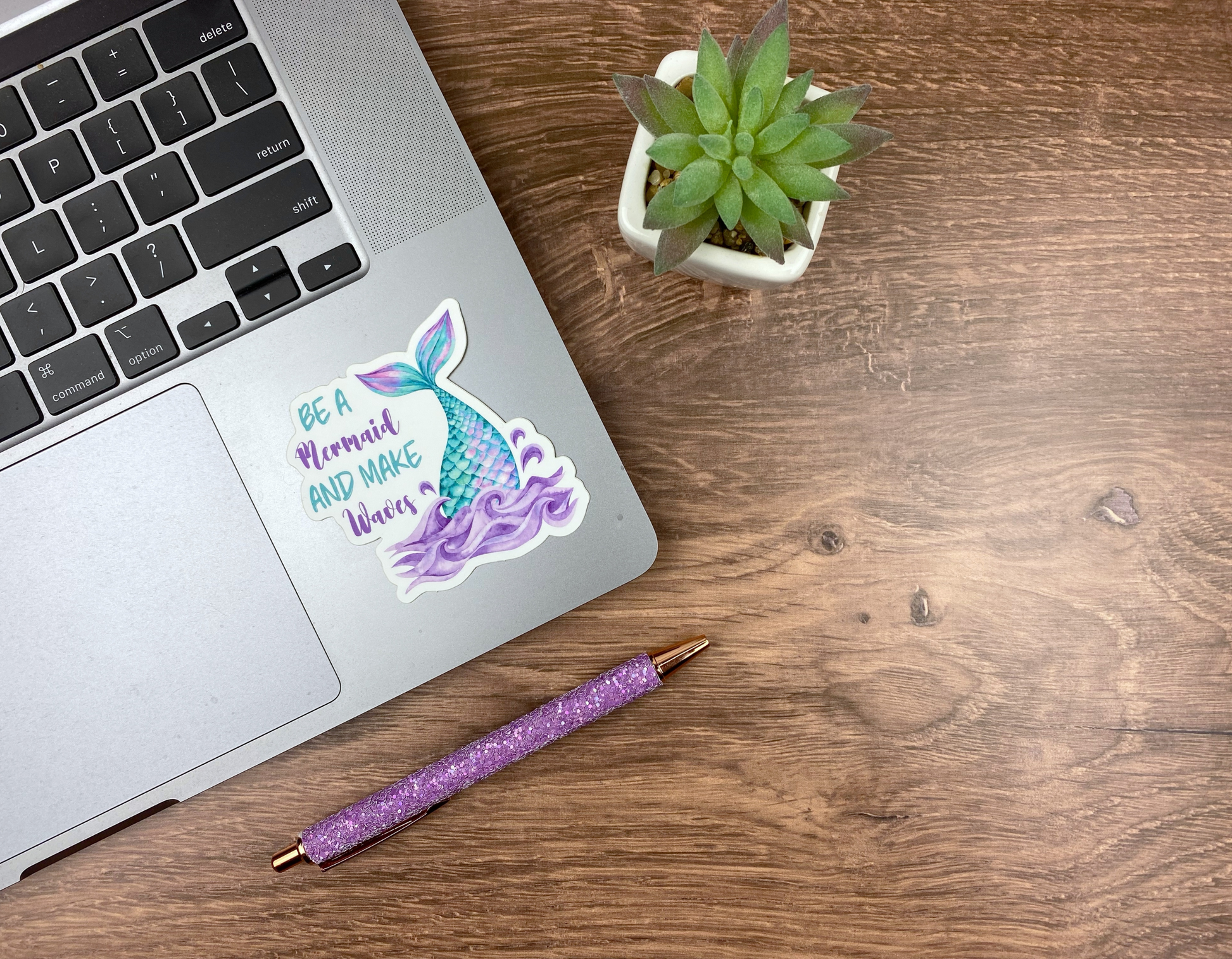 Mermaid tail and waves sticker applied to the corner of a silver laptop trackpad, next to a succulent and a glitter pen on a wood desk