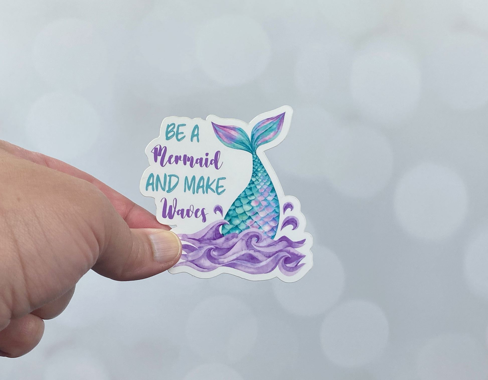 Die-cut vinyl sticker held between fingers, showing a teal and purple mermaid tail rising out of purple waves, with the text "Be a mermaid and make waves," on a light bokeh background