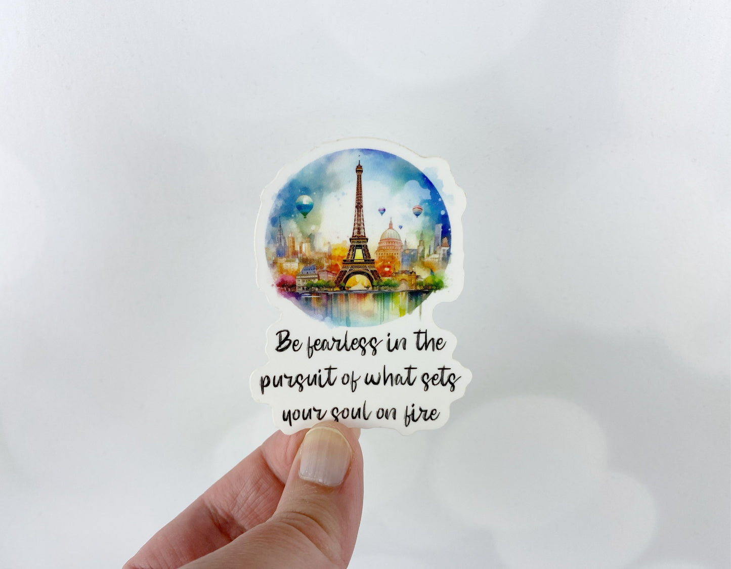 A hand holds a sticker illustration of a watercolor skyline with the Eiffel Tower and hot air balloons, reading "Be fearless in the pursuit of what sets your soul on fire," against a white background.