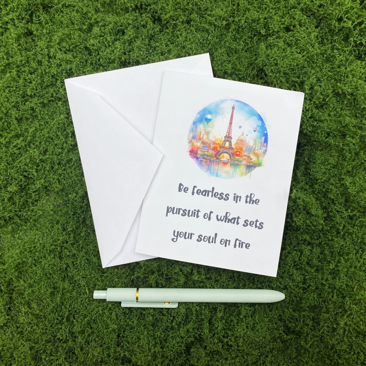 Folded greeting card featuring a watercolor Paris skyline and the quote "Be fearless in the pursuit of what sets your soul on fire," resting on a white envelope on a mossy surface, with a sage green pen below.