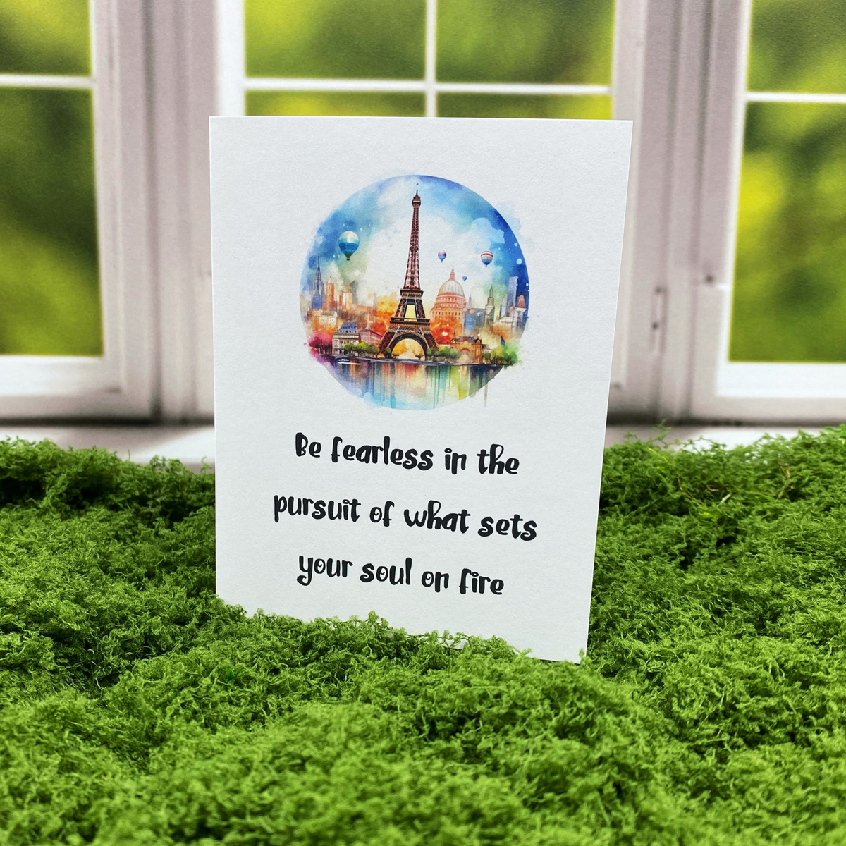 Greeting card with a watercolor illustration of the Eiffel Tower and Paris skyline in a circular frame, with hot air balloons in the sky, above the text "Be fearless in the pursuit of what sets your soul on fire," standing upright on a moss surface in front of a white-framed window.