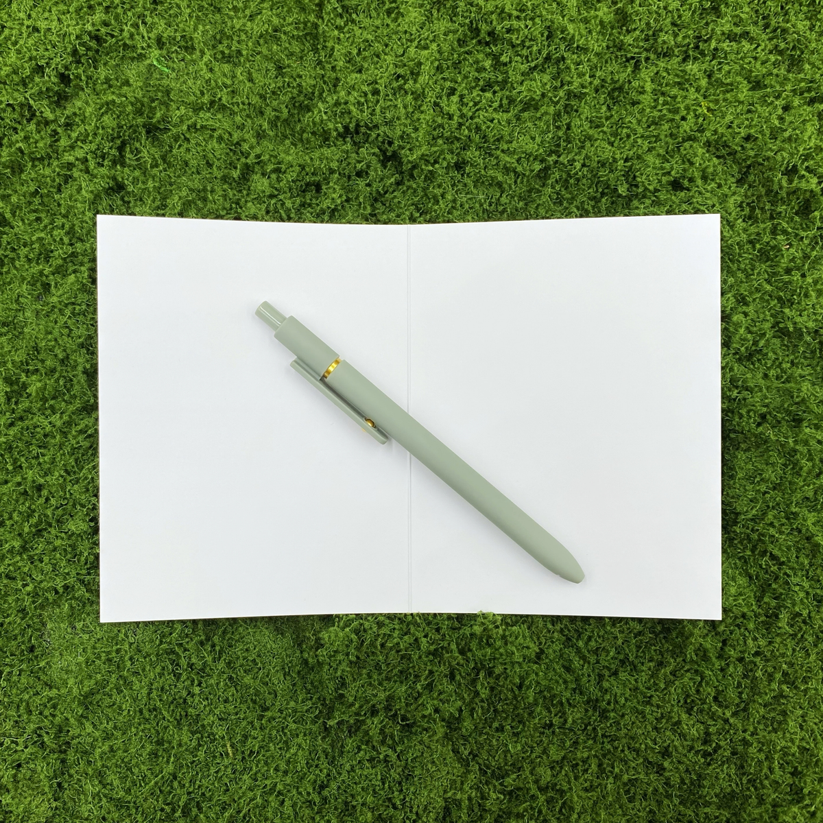 Card opened to show the blank white interior, ready for a handwritten message, with a sage green pen laid across the crease on a mossy background.