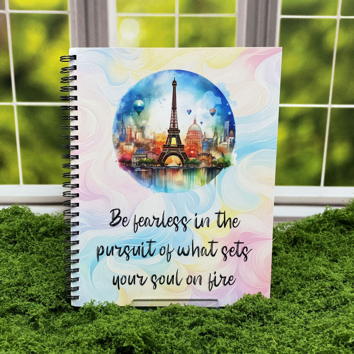 Spiral-bound notebook with a pastel swirl cover and a circular watercolor illustration of the Paris skyline with the Eiffel Tower and hot air balloons, reading 'Be fearless in the pursuit of what sets your soul on fire,' displayed on a moss mat with a window backdrop.