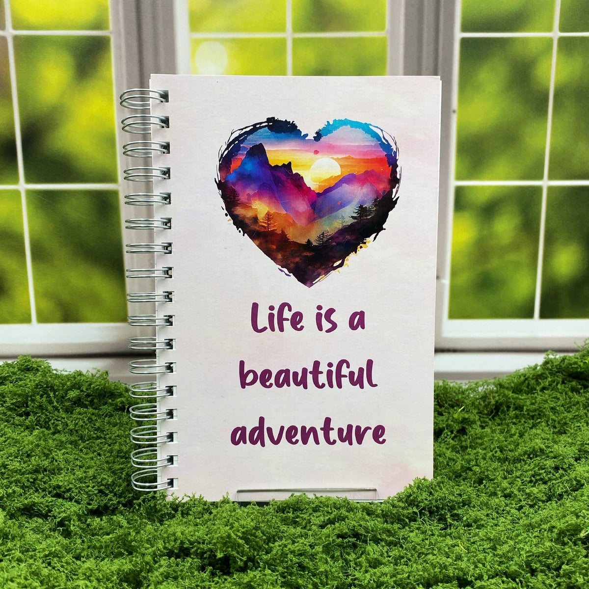 Spiral-bound white notebook with a watercolor heart-shaped sunset mountain illustration above text reading 'Life is a beautiful adventure,' displayed on a moss mat with a window backdrop.