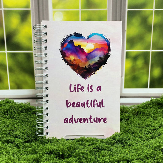 Spiral-bound white notebook with a watercolor heart-shaped sunset mountain illustration above text reading 'Life is a beautiful adventure,' displayed on a moss mat with a window backdrop.