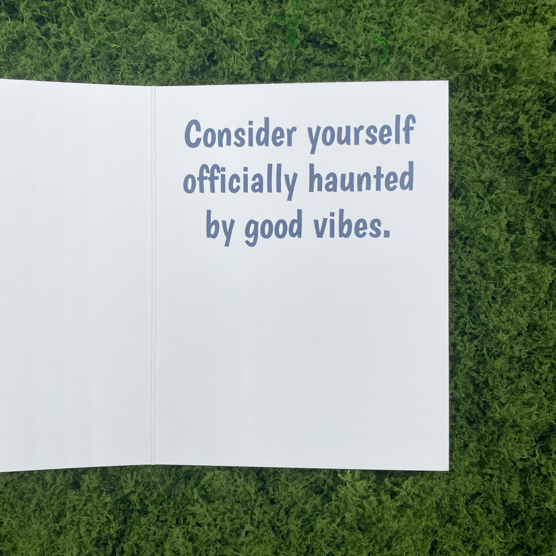 Inside spread of the greeting card showing the printed message "Consider yourself officially haunted by good vibes." lying open on artificial grass.