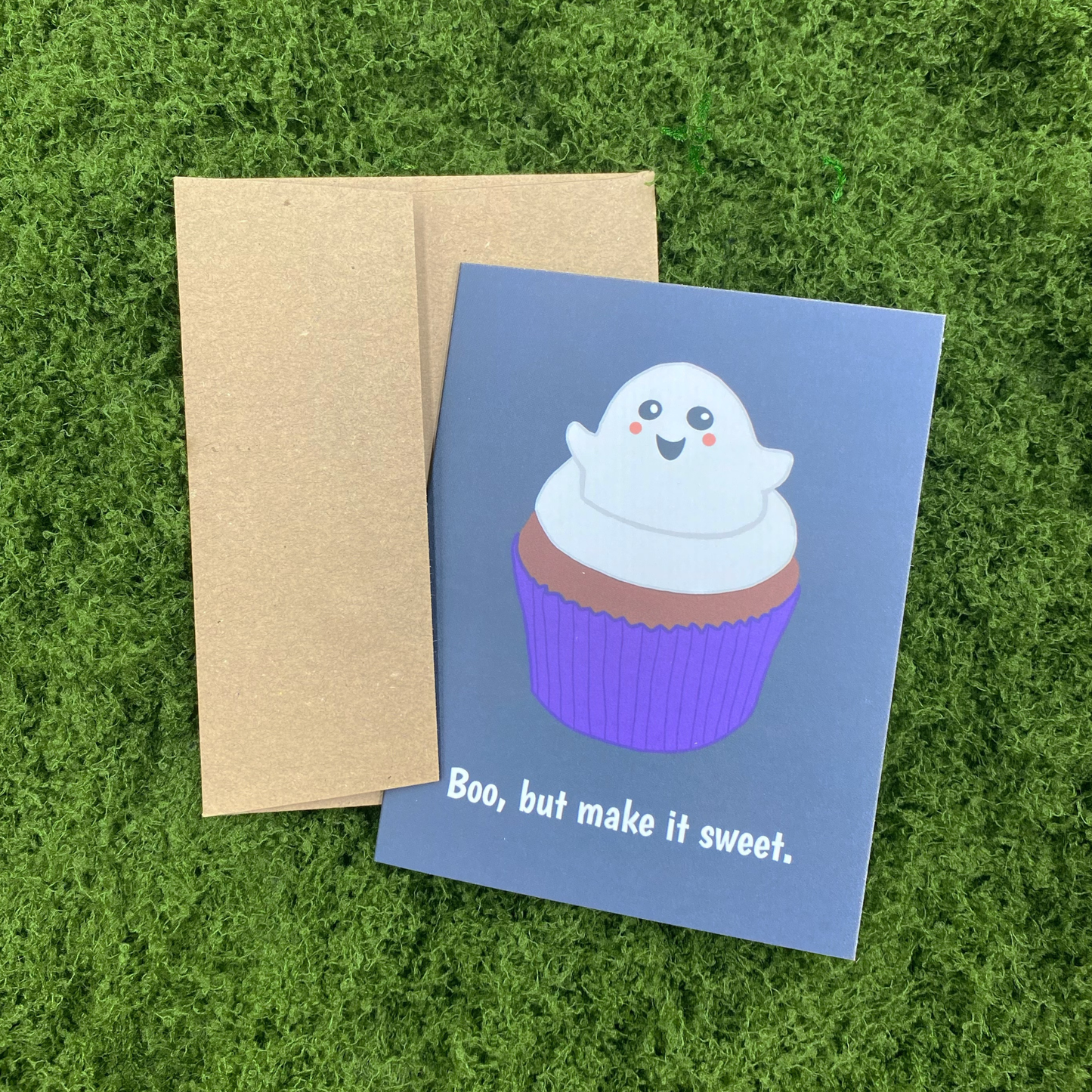 Greeting card that reads "Boo, but make it sweet." with an illustration of a smiling ghost-shaped frosting swirl atop a chocolate cupcake with purple liner, resting against its Kraft paper envelope on artificial grass.