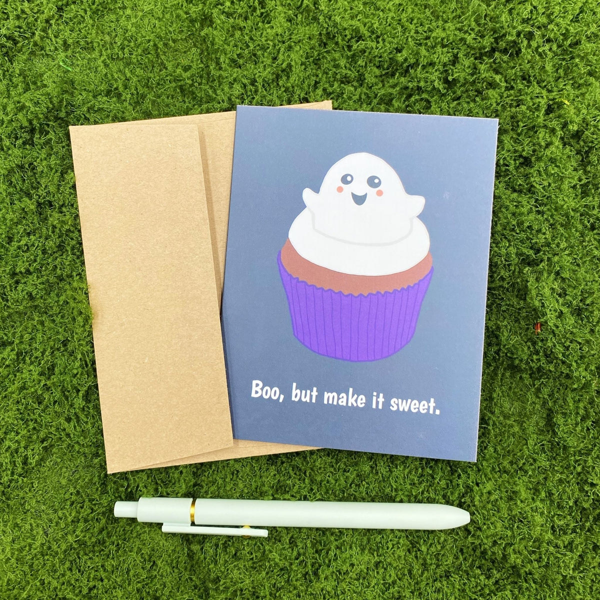 "Boo, but make it sweet." greeting card lying on a kraft brown envelope on grass, with a mint green pen beside it for scale.