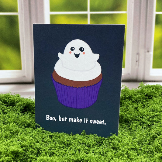 "Boo, but make it sweet." greeting card standing upright on a moss mat in front of a window backdrop, featuring an illustrated smiling ghost as frosting atop a chocolate cupcake with a purple wrapper.