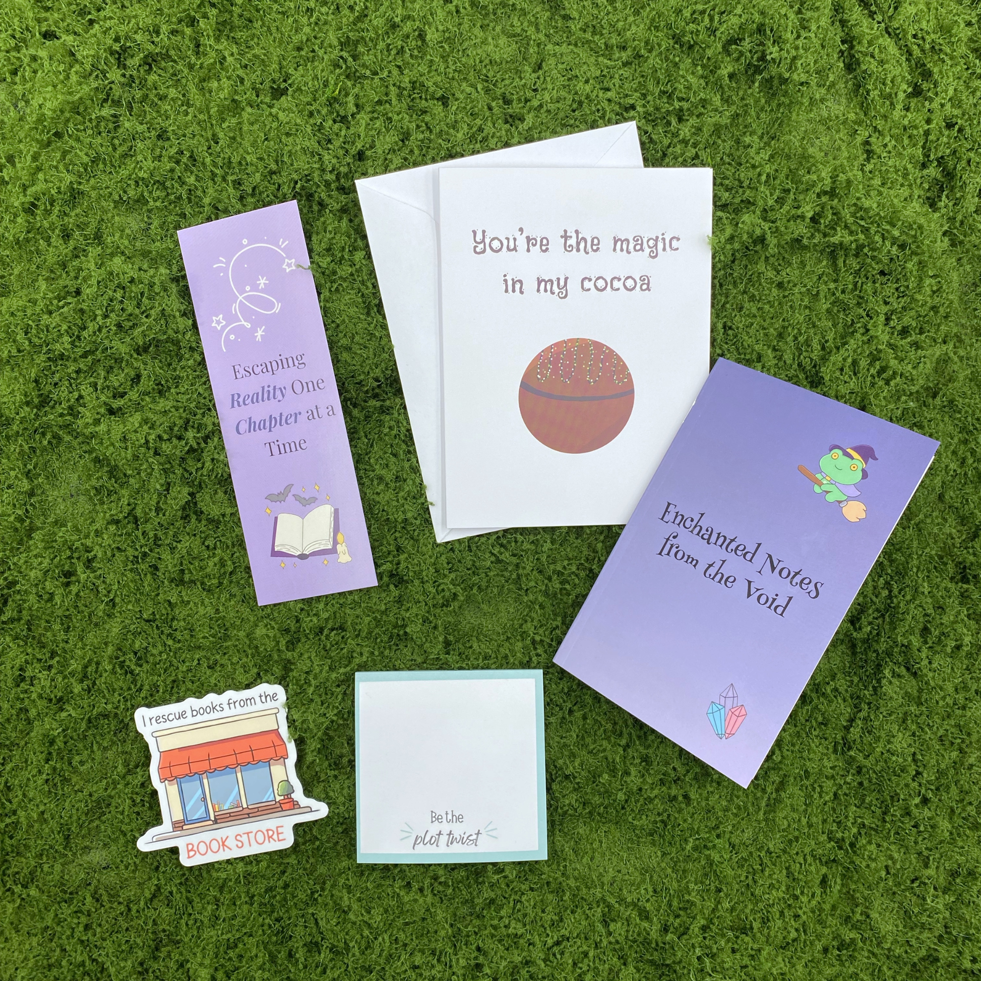 A gift set including a purple bookmark, a you're the magic in my cocoa greeting card with envelope, an enchanted notes pocket notebook, a be the plot twist sticky note pad, and a bookshop die-cut sticker, arranged on a green moss surface