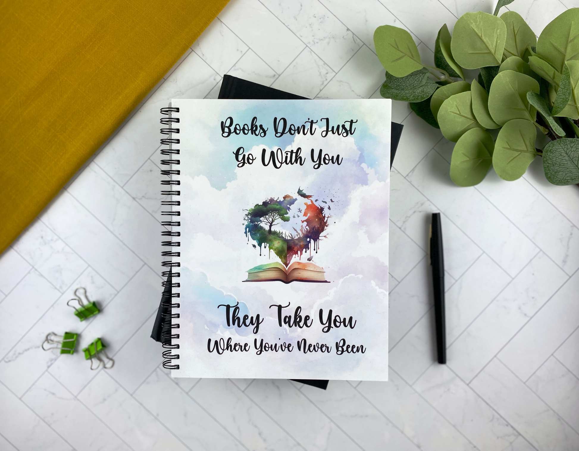 Notebook cover with a soft cloud-and-sky watercolor background, an open book with a heart-shaped tree and landscape rising from its pages, and the words "Books don't just go with you, they take you where you've never been," resting on a black notebook with green binder clips and a black pen nearby.