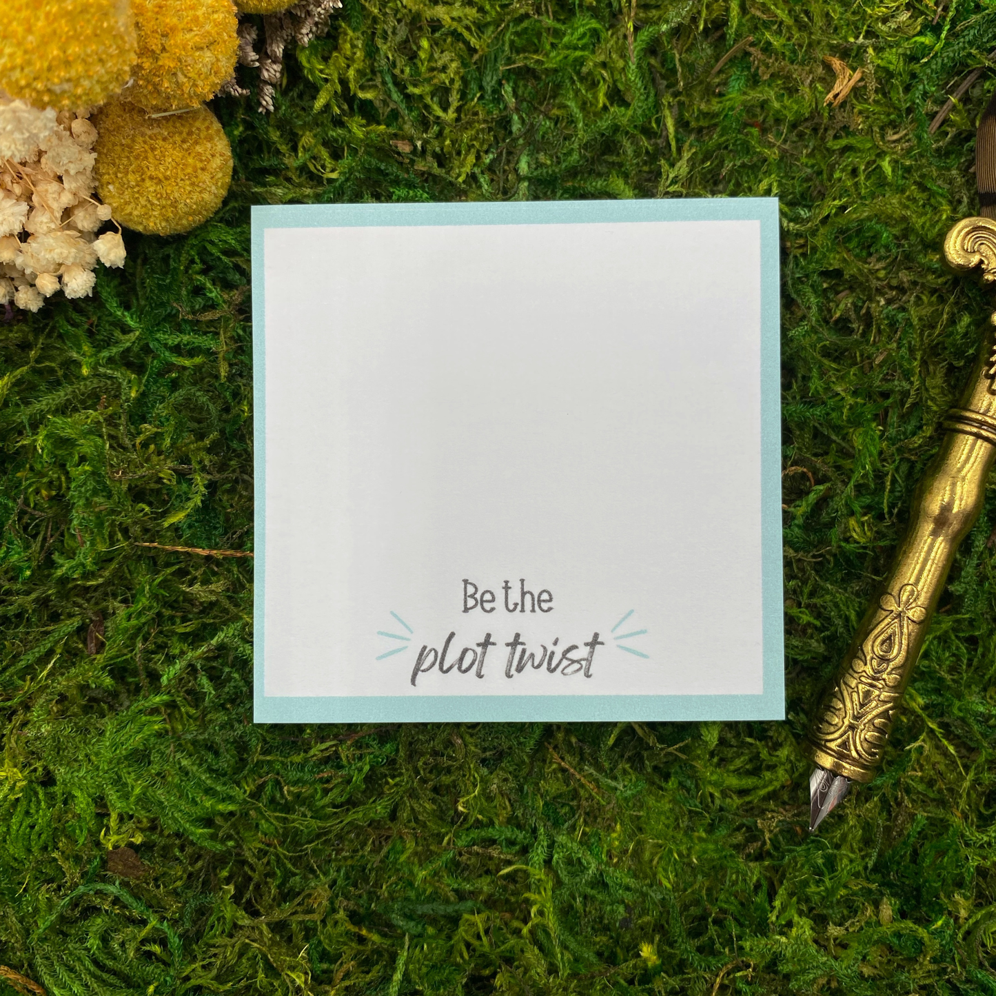 Square sticky note pad with a teal border reading "Be the plot twist" in black script, resting on moss next to dried yellow and white flowers and a gold quill pen.