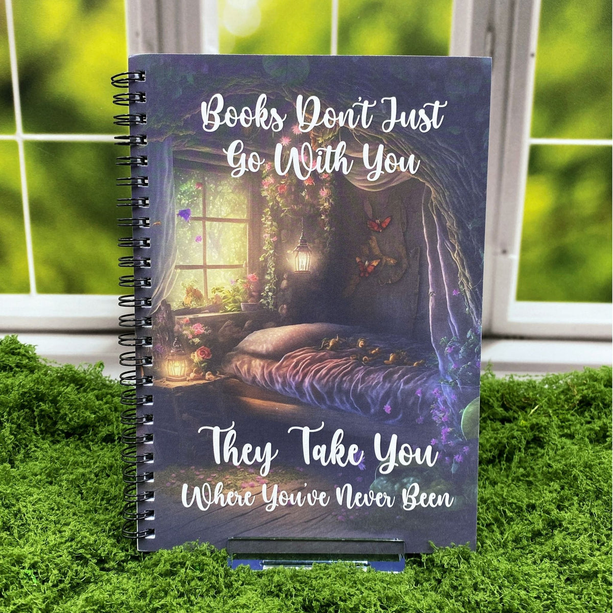 Spiral-bound notebook with an illustrated fairycore reading nook featuring a canopy bed, glowing lanterns, and butterflies, reading 'Books don't just go with you. They take you where you've never been,' displayed on a moss mat with a window backdrop.