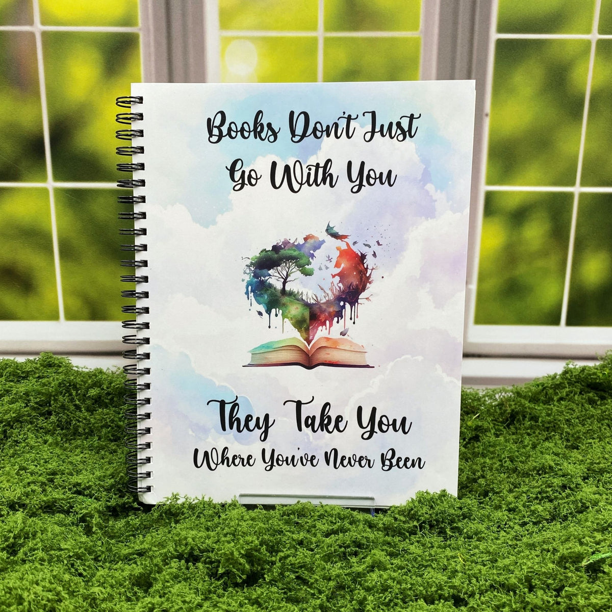 Spiral-bound notebook with a cloudy watercolor sky background and a heart-shaped illustration of a tree and cosmic sky rising from an open book, reading 'Books don't just go with you. They take you where you've never been,' displayed on a moss mat with a window backdrop.