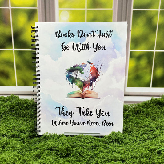 Spiral-bound notebook with a cloudy watercolor sky background and a heart-shaped illustration of a tree and cosmic sky rising from an open book, reading 'Books don't just go with you. They take you where you've never been,' displayed on a moss mat with a window backdrop.