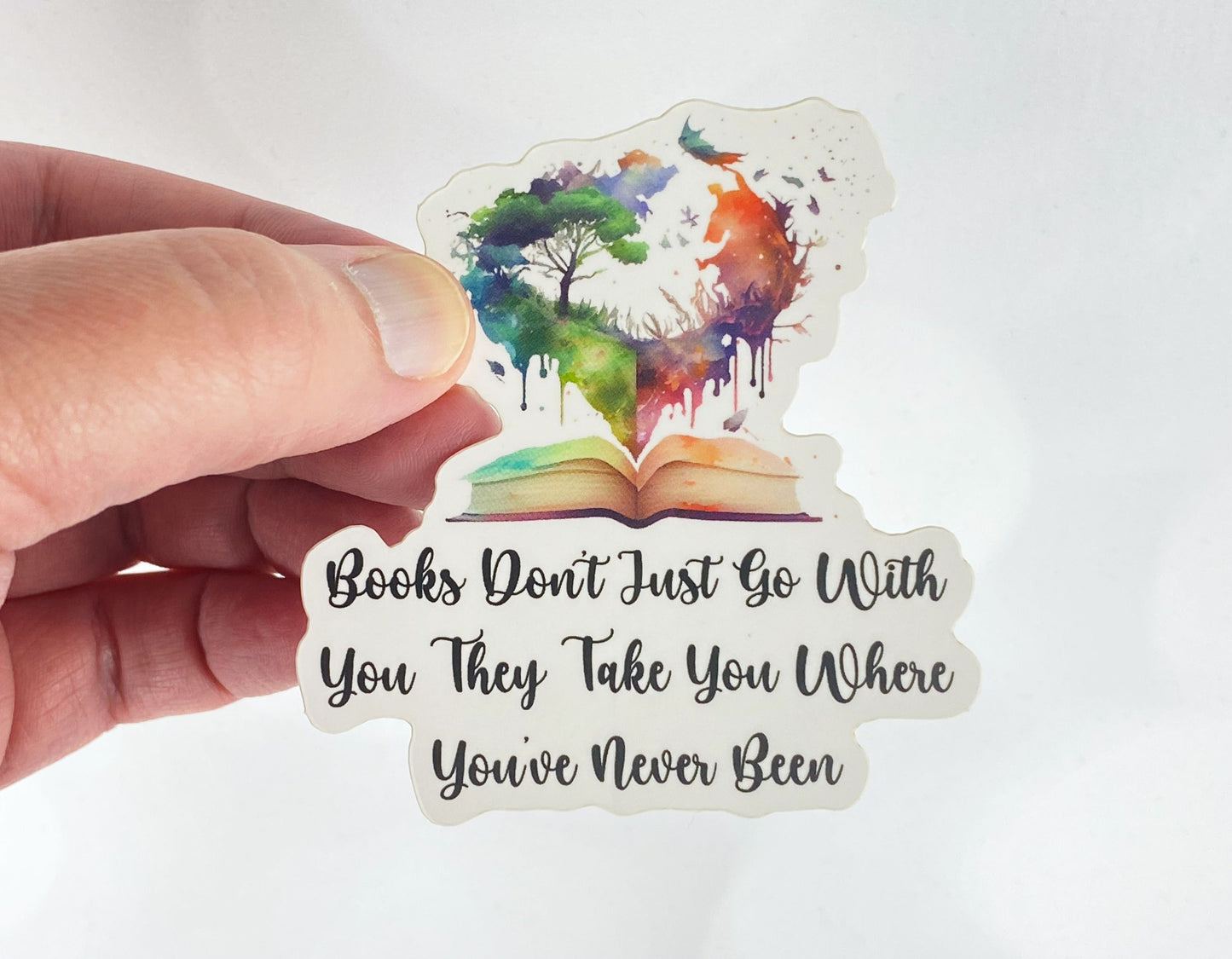 A hand holds a sticker illustration of a watercolor tree and galaxy rising from an open book, reading "Books don't just go with you, they take you where you've never been," against a white background.
